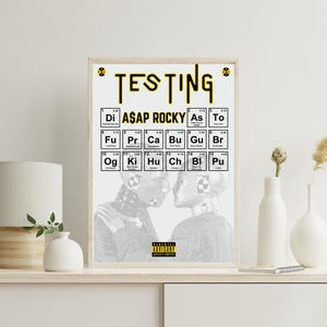 Puede incluir: Una impresión artística enmarcada con la palabra "TESTING" en amarillo, encima de un diseño de estilo tabla periódica con "ASAP ROCKY". La impresión incluye una imagen en blanco y negro de dos figuras. El marco es marrón claro.