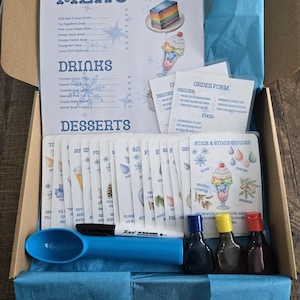 May include: An open cardboard box filled with blue tissue paper, containing a menu, order forms, and dessert cards. Includes a blue scoop, a black marker, and small bottles of colored liquids. The menu lists drinks and desserts.