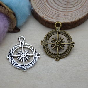 Puede incluir: Dos dijes de brújula de plata y bronce con un diseño de estrella y las letras N, S, E y W alrededor del borde.