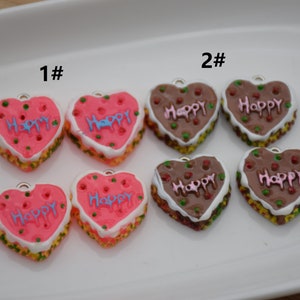 Può includere: Due set di biscotti a forma di cuore con la scritta "Happy" su di essi. Il primo set è rosa con la scritta blu e il secondo set è marrone con la scritta rosa.