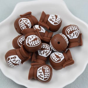 May include: Brown beads with the text "Pot Head" in white lettering. The beads are shaped like a pot and are arranged on a white, scalloped plate. The background is a light blue.