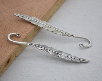 May include: Two silver feather-shaped bookmarks with a curled hook at the end. The bookmarks are on a wooden surface.