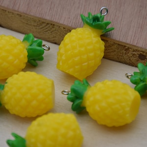 Puede incluir: Un grupo de seis abalorios de piña amarillos con hojas verdes y anillos de salto plateados. Los abalorios son de plástico y son perfectos para añadir un toque tropical a las joyas o las manualidades.
