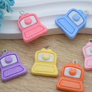 Puede incluir: Una colección de coloridos dijes de ordenador portátil en rosa, azul, morado, amarillo y naranja. Cada dije presenta una pantalla blanca con un diseño en forma de gato y un teclado. Los dijes tienen un lazo plateado para sujetarlos a joyas u otras manualidades.