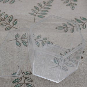 Puede incluir: Una caja hexagonal de plástico transparente con tapa. La caja está vacía y se encuentra sobre una superficie de tela con un patrón floral.