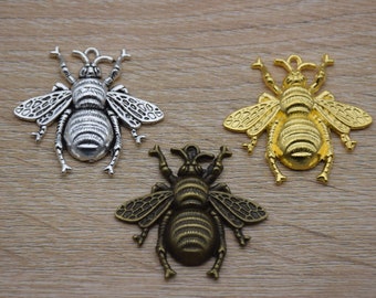 May include: Three metal bee-shaped embellishments in silver, gold, and bronze tones. The bees have detailed wings and bodies, with antennae raised. The items are displayed on a light brown wooden surface.