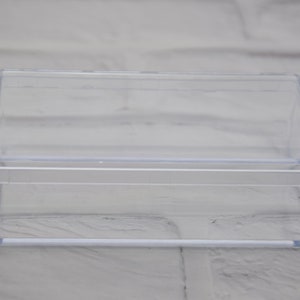 Könnte beinhalten: Eine transparente Plastik-Aufbewahrungsbox mit einem Scharnierdeckel. Die Box ist leer und hat eine rechteckige Form.