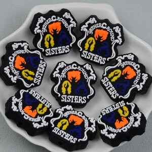 以下が含まれることがあります： 白い縁取りと「GATHER ROUND SISTERS」の文字が施された、7つの黒いシリコン製チャーム。各チャームには、鮮やかなオレンジ、黄色、紫色の3つの様式化された髪のシルエットが円形に配置されています。