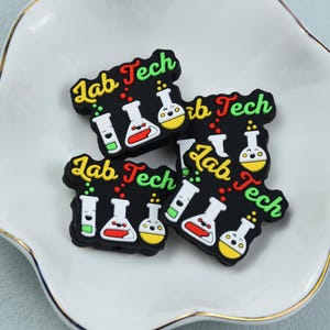 Puede incluir: Varios amuletos de goma negra con la inscripción "Lab Tech" en letras amarillas, rojas y verdes. Cada amuleto presenta ilustraciones de dibujos animados de equipos de laboratorio, incluyendo vasos de precipitados y matraces, en blanco, rojo y amarillo. Los amuletos están colocados sobre un plato blanco festoneado con borde dorado.