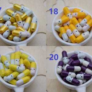 Puede incluir: Una colecci&oacute;n de cuentas coloridas en forma de c&aacute;psula sonriente en peque&ntilde;os cuencos blancos. Las cuentas est&aacute;n en varias combinaciones de colores, incluyendo oro y blanco, amarillo y blanco, y morado y blanco. Los n&uacute;meros 17, 18, 19 y 20 son visibles.