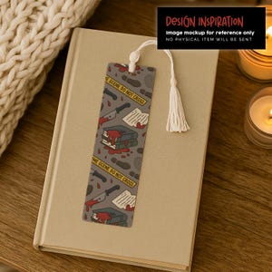 May include: A gray bookmark with a white tassel, featuring a crime scene design. The design includes knives, books, footprints, and the text "CRIME SCENE DO NOT CROSS". The bookmark is on a beige book.