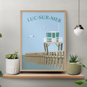 Peut inclure: Une affiche encadrée représentant une scène côtière avec une tour de sauveteur et une jetée. L'affiche a un ciel bleu clair avec le texte "LUC-SUR-MER" en haut. Deux mouettes volent. Le cadre est en bois.
