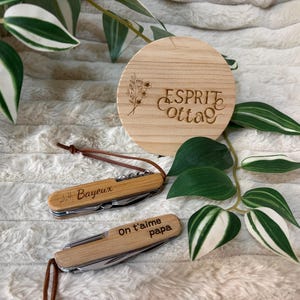 Op de afbeelding: Twee houten zakmessen met bruine leren koorden, één gegraveerd met "Bayeux" en de andere met "On t'aime papa". Een ronde houten onderzetter met "Esprit Cottage" en een bloemmotief is ook aanwezig. Groene en witte bladeren zijn op de achtergrond.