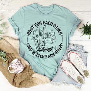 May include: A light blue t-shirt with a black cactus graphic and the text "Root for each other and watch each grow." The shirt is paired with denim shorts, white sneakers, and a mug with heart designs.