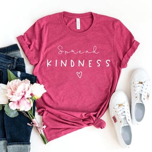 May include: A magenta t-shirt with the words "Spread KINDNESS" in white script and a heart symbol. The shirt is paired with blue jeans, white sneakers, and a bouquet of pink and white flowers. The shirt is folded at the bottom.