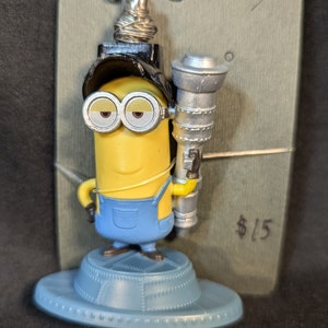 May include: A yellow Minion figurine wearing blue overalls, a black hat, and goggles, holding a silver ray gun. The figurine stands on a light blue, saucer-shaped base. The Minion has a silver antenna on its head.