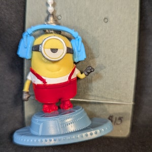May include: A yellow Minion figure wearing blue headphones and a red jumpsuit, standing on a gray spaceship-shaped base. The figure is holding a microphone in its right hand. The base has a $15 price tag.