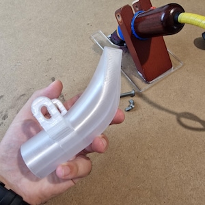 May include: A clear, curved plastic component with a loop attachment. The object is held in a hand, with a wooden and metal apparatus in the background. A bolt and nut are also visible.