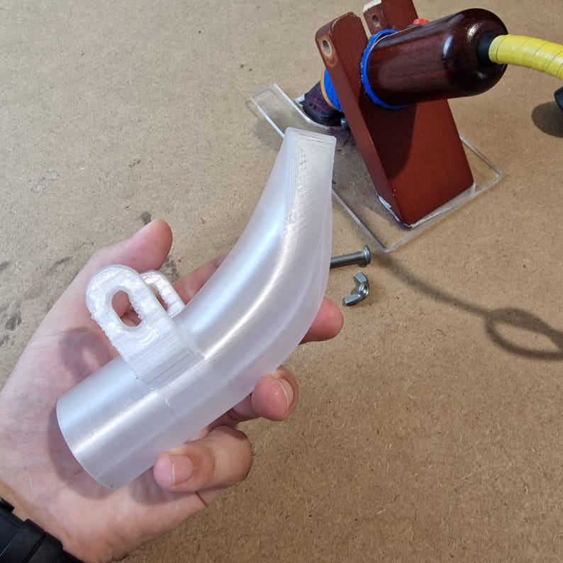 May include: A clear, curved plastic component with a loop attachment. The object is held in a hand, with a wooden and metal apparatus in the background. A bolt and nut are also visible.