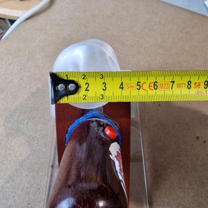 May include: A brown bottle with a red and white design, measured with a yellow tape measure. The bottle is approximately 2 inches wide. A clear plastic piece is on top of the bottle.