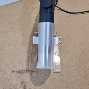 May include: A clear, cylindrical plastic holder with a black device inserted. The holder is attached to a clear plastic base with a rectangular shape. The setup is on a brown surface.