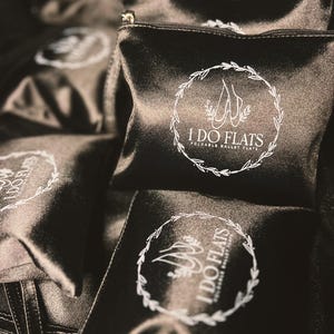 May include: Black satin pouches with a zipper closure. Each pouch features a white circular logo with the text "I DO FLATS" and a ballet shoe design. The pouches are designed to hold foldable ballet flats.