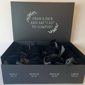 May include: A large black rectangular box filled with multiple pairs of black slippers. The interior lid features white text reading GRAB A PAIR AND SAY I DO TO COMFORT. The front of the box displays size labels: SMALL (6-7), MEDIUM (8-9), and LARGE (10-11).