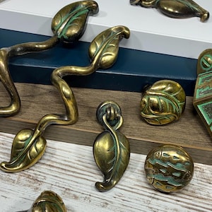 Peut inclure: Ensemble de quincaillerie de meubles en laiton antique avec des motifs de feuilles et géométriques. L'ensemble comprend des poignées, des boutons et des tirettes avec une finition vieillie, dans des tons or et vert. Idéal pour ajouter une touche vintage.