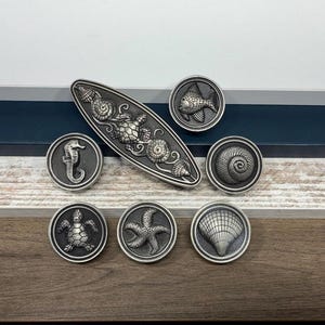 May include: A collection of seven silver-toned metal cabinet knobs featuring nautical motifs. The set includes one elongated oval knob with a sea turtle and shells, and six circular knobs depicting a seahorse, fish, spiral shell, sea turtle, starfish, and scallop shell.