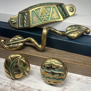 Op de afbeelding: Een verzameling antieke messing kastbeslagen. Inclusief een lange handgreep met geometrische ontwerpen en twee blad-vormige knoppen. De hardware heeft een verweerde afwerking met hints van groene patina, wat duidt op leeftijd en gebruik.