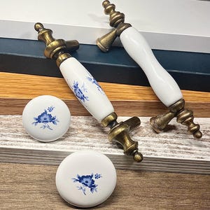 Pode incluir: Duas puxadores de armário brancos e dourados com um design floral e dois botões redondos a condizer. As puxadores têm um centro de cerâmica branca com padrões florais azuis e detalhes em metal dourado. Os botões são brancos com um design floral azul.