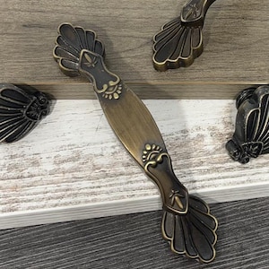 May include: Antique bronze cabinet pulls with a decorative fan-shaped design. The pulls have an oval center with ornate detailing and are set against a light wood background. The hardware is ideal for adding a vintage touch to furniture.