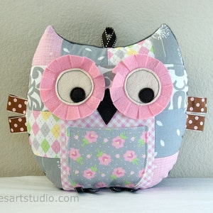 May include: A patchwork owl-shaped pillow in pink, gray, and white fabrics. The owl has large pink felt eyes with black pupils, a black beak, and a small pocket with a floral pattern. Brown polka dot ribbons are attached to the side.