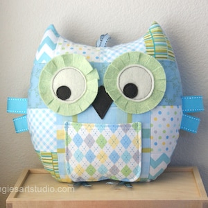 May include: A patchwork owl-shaped pillow in shades of blue, green, and white. The owl features large, round eyes with green felt rims and black pupils. Blue ribbon accents and a small pocket add detail. The pillow is made of various patterned fabrics.