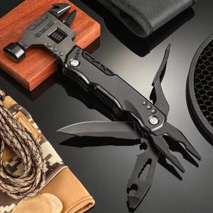 Op de afbeelding: Een zwarte multitool met een hamer, verstelbare moersleutel, tang, mes en andere gereedschappen. De multitool ligt op een houten blok en een reflecterend oppervlak. Een bruine handschoen en een zwart zakje zijn ook zichtbaar.