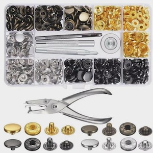 Puede incluir: Un organizador de plástico transparente lleno de varios cierres de presión metálicos en plata, oro y negro. Incluye herramientas para colocar broches y una herramienta plateada para colocar broches. Se muestra una variedad de estilos de broches debajo.