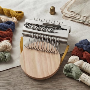 May include: A metal darning loom with a circular wooden base, featuring a row of vertical metal hooks and a sliding metal bar. The tool is surrounded by various small skeins of colored yarn in shades of mustard, blue, rust, cream, and sage green.