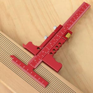 May include: A red T-square and ruler tool with inch measurements. The tool is made of metal and has a sliding mechanism. The tool is used for woodworking and measuring. The tool is on a wooden surface.