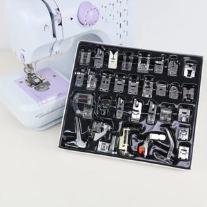 May include: A white and lavender mini sewing machine with a set of various presser feet. The presser feet are arranged in a black tray. The sewing machine has a dial and a needle plate.