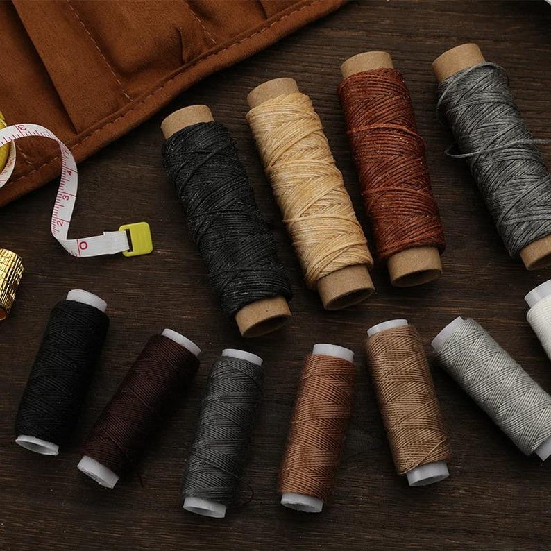 May include: A collection of spools of thread in various colors, including black, brown, gray, and beige. The thread is wound around cardboard tubes. A measuring tape and thimble are also visible.