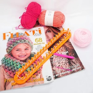 Puede incluir: Un telar de punto naranja con hilo blanco, un ganchillo rosa y tres ovillos de lana rosa y melocotón. El telar está colocado sobre una revista con el texto "FIATI Kids" y una foto de un niño con gorro y bufanda de punto.