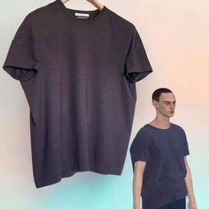 May include: A dark purple, short-sleeved t-shirt hangs on a wooden hanger. A mannequin wears a similar dark purple t-shirt and dark blue jeans. The mannequin is wearing black sneakers with a colorful design.