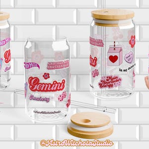 Puede incluir: Vasos de vidrio transparente con tapas y pajitas de bambú. Los vasos están decorados con pegatinas rosas y rojas con cerezas, flores y la palabra "Gemini". Una persona sostiene un vaso. El fondo es una pared de azulejos blancos.