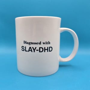 May include: White ceramic mug with a curved handle. The mug features the text "Diagnosed with SLAY-DHD" in black, centered on the front. The mug is set against a solid blue background.