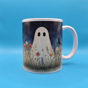 White ceramic mug with a whimsical design of a ghost standing in a meadow of wildflowers under a night sky with stars, witches and a bat. Cozy, cute, and spooky aesthetic, perfect for Halloween, cottagecore vibes, and autumn coffee moments.