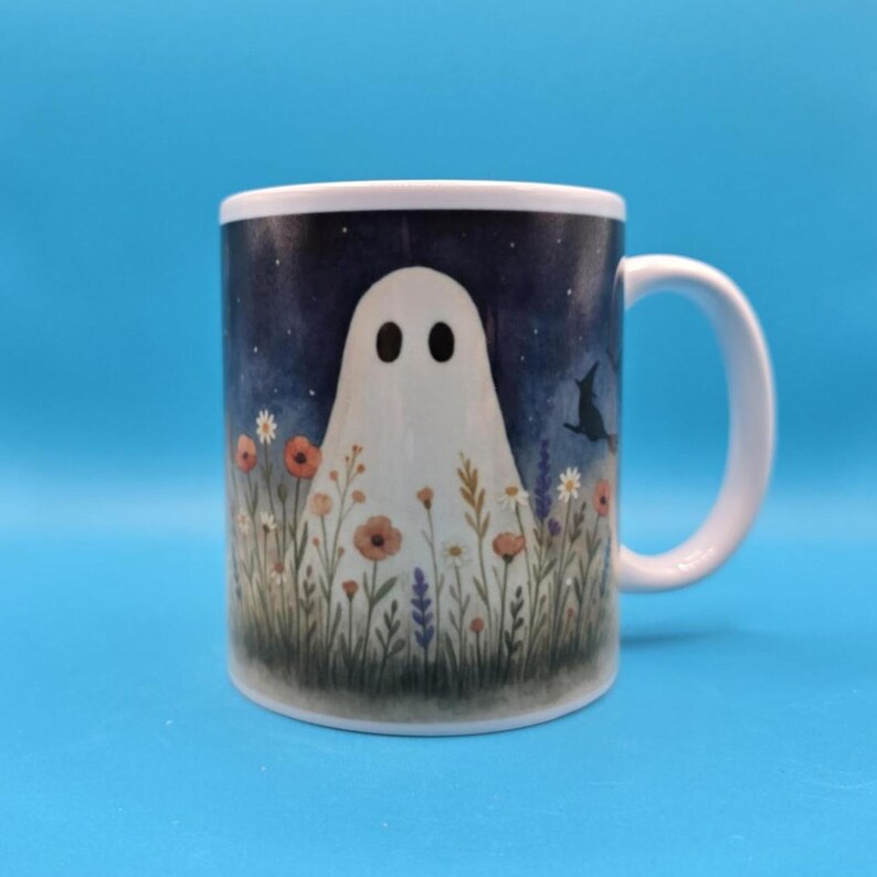 White ceramic mug with a whimsical design of a ghost standing in a meadow of wildflowers under a night sky with stars, witches and a bat. Cozy, cute, and spooky aesthetic, perfect for Halloween, cottagecore vibes, and autumn coffee moments.
