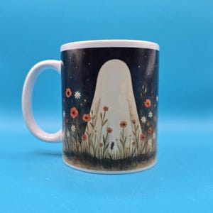 White ceramic mug with a whimsical design of a ghost standing in a meadow of wildflowers under a night sky with stars, witches and a bat. Cozy, cute, and spooky aesthetic, perfect for Halloween, cottagecore vibes, and autumn coffee moments.