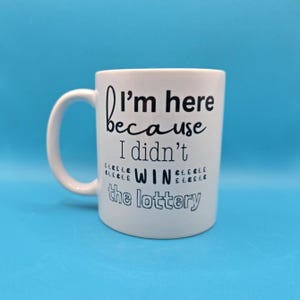 May include: White ceramic mug with a curved handle. The mug features the text "I'm here because I didn't win the lottery" in black lettering. The mug is set against a solid blue background.