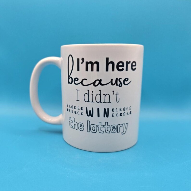 May include: White ceramic mug with a curved handle. The mug features the text "I'm here because I didn't win the lottery" in black lettering. The mug is set against a solid blue background.