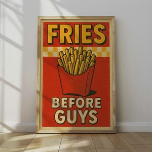 May include: A framed poster with a retro design. The poster features the words "FRIES BEFORE GUYS" in yellow and white text. A red box of fries is in the center. The background is red with a yellow and white checkered pattern.